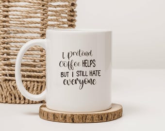 Puede incluir: Una taza de cerámica blanca con asa que presenta el texto impreso en negro I pretend coffee HELPS BUT I STILL HATE everyone en una mezcla de letras cursivas y de imprenta. La taza está colocada sobre un posavasos de madera con corteza.