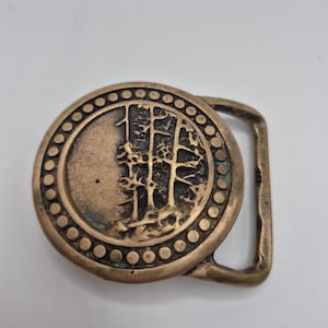May include: A round brass belt buckle featuring a raised relief design of trees on one side and a smooth surface on the other. The circular center is framed by a border of small, evenly spaced raised dots, with a rectangular loop on the right side.