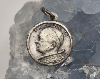 May include: A circular silver-toned metal pendant featuring a profile relief of Pope Paul VI facing left. The text PAULUS VI is embossed along the upper edge of the coin-like charm, which includes a small metal loop at the top for attachment.