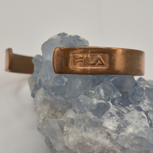 May include: A copper-colored cuff bracelet with the FILA logo embossed on the front. The bracelet is resting on a light blue crystal formation. The cuff is a simple, minimalist design.