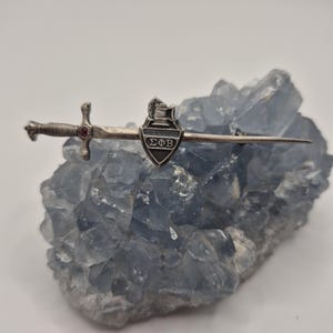 May include: A silver-toned brooch featuring a sword design with a shield emblem. The sword has a red gemstone at the hilt. The shield has the Greek letters ΣΦΒ. The brooch is displayed on a blue crystal.