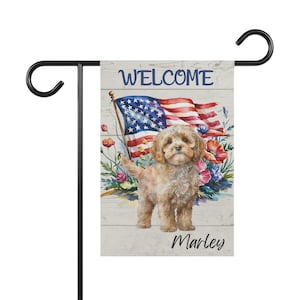 May include: A garden flag with a tan dog, an American flag, and floral details. The flag has the word "WELCOME" at the top and "Marley" at the bottom. It's mounted on a black metal stand. The flag's design is on a white wooden background.