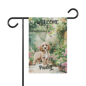 Puede incluir: Bandera de jardín con una ilustración de un Cocker Spaniel en un jardín. La palabra "WELCOME" está en la parte superior y "Pauley" en la inferior. La bandera está montada en un soporte de metal negro. La imagen incluye flores y un camino de piedra.