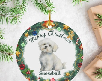 Puede incluir: Adorno navideño redondo con un perro maltés blanco llamado Snowball. El adorno tiene un diseño de corona verde con detalles en rojo, azul y dorado, y las palabras "Merry Christmas" están escritas encima del perro.
