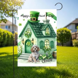 Puede incluir: Una bandera de jardín del Día de San Patricio con una casa verde con un sombrero de duende, tréboles y el texto "Happy St. Patrick's Day". Un perro llamado Charlie está sentado frente a la casa. La bandera está en un soporte de metal negro.