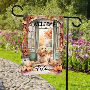 May include: A garden flag with a watercolour illustration of a dog, pumpkins, and autumn leaves. The flag reads "WELCOME" above the dog and "Pixie" below. The flag is displayed on a black metal stand in a garden.
