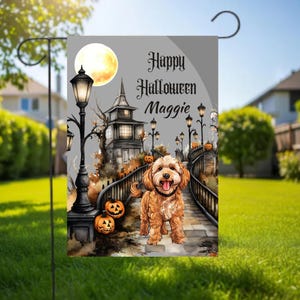 May include: A rectangular garden flag featuring a golden-brown dog standing on a stone bridge. The scene includes a haunted house, glowing jack-o'-lanterns, and black street lamps under a full moon. The text reads Happy Halloween Maggie.