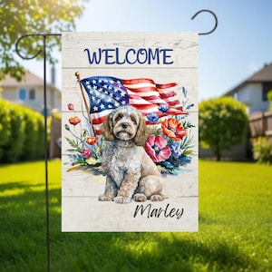 Puede incluir: Una bandera de jardín con un Cavalier King Charles Spaniel, con pelaje marrón y blanco, frente a una bandera estadounidense y flores. La palabra "WELCOME" está en la parte superior y el nombre "Marley" en la parte inferior. Fondo de madera.