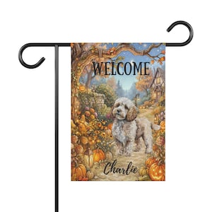 May include: A rectangular garden flag hanging on a black metal stand, featuring an illustration of a light-coloured dog standing on a path surrounded by orange pumpkins, autumn foliage, a stone wall, and a house, with the text WELCOME and Charlie.