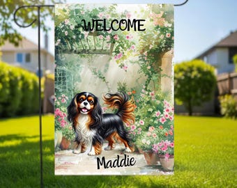 May include: A garden flag featuring a Cavalier King Charles Spaniel with a black, white, and brown coat, standing in front of a floral backdrop. The flag reads "WELCOME" at the top and "Maddie" at the bottom.