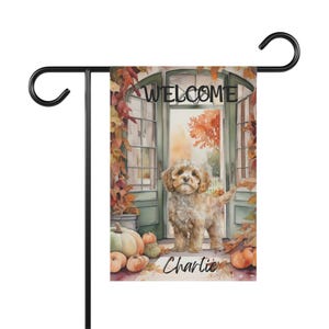 Puede incluir: Una bandera de jardín rectangular colgada en un soporte de metal negro. La bandera presenta una ilustración en acuarela de un perro de pelo rizado color marrón claro en una puerta abierta con hojas de otoño, calabazas y el texto WELCOME y Charlie.