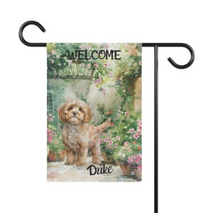 May include: A garden flag with a watercolour illustration of a light brown dog named Duke. The flag reads "WELCOME" above the dog, set against a backdrop of flowers and a stone wall. The flag is attached to a black metal stand.