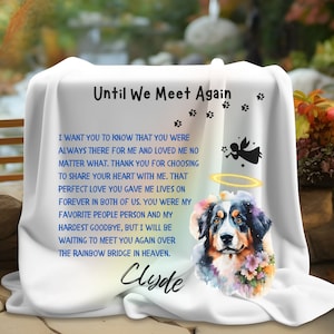 Puede incluir: Manta conmemorativa blanca con una pintura de acuarela de un perro de montaña bernés con un halo. La manta tiene el texto "Until We Meet Again" y "Clyde" en texto azul.