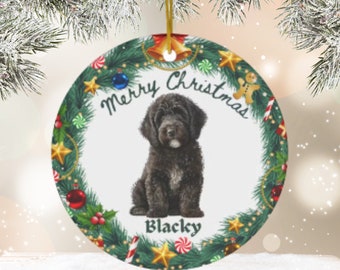 May include: A round Christmas bauble featuring a green wreath border. The wreath is decorated with red and green baubles, candy canes, and stars. In the centre is a black dog with the text "Merry Christmas" and "Blacky" below the dog.