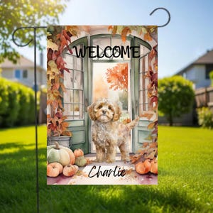 Pode incluir: Uma bandeira de jardim retangular com uma ilustração em aquarela de um filhote de cachorro marrom claro em pé em uma porta aberta. O texto "WELCOME" aparece no topo e o nome "Charlie" está escrito na parte inferior. Folhas de outono e abóboras cercam a porta.