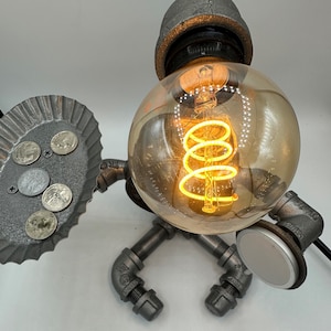 Op de afbeelding: Een unieke lamp in industriële stijl gemaakt van metalen buizen en fittingen. De lamp heeft een grote, ronde gloeilamp met een warme, gloeiende gloeidraad. Een klein dienblad bevat verschillende munten. Het ontwerp van de lamp doet denken aan een robot.