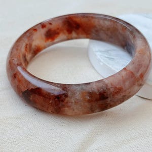 May include: A polished, circular bangle bracelet crafted from translucent stone featuring a marbled pattern of deep reddish-brown, rust, and clear quartz-like inclusions. The smooth, rounded surface displays natural variations in color and texture.