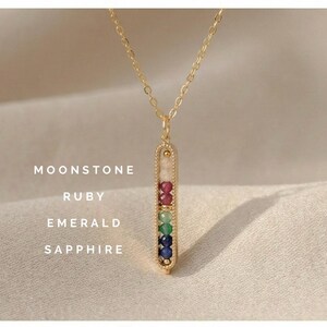 Puede incluir: Un collar dorado con un colgante vertical que contiene piedras preciosas de luna, rubí, esmeralda y zafiro. El collar se muestra sobre una tela de tono neutro, con las piedras dispuestas en una línea vertical colorida.