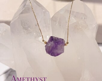 May include: A delicate gold chain necklace with a raw amethyst crystal pendant. The amethyst is a deep purple color and has a natural, rough texture. The necklace is displayed on a white crystal background.