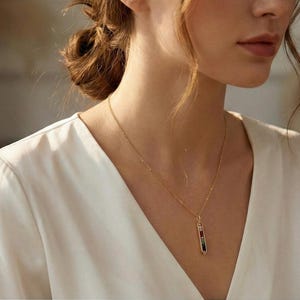 May include: A gold necklace with a pendant featuring red, black, and green accents. The pendant is a vertical rectangle shape. The necklace is worn over a white v-neck top.