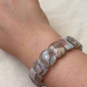 May include: A stretch bracelet composed of smooth, polished, rectangular-shaped beads made of translucent gray stone with internal white, pink, and orange inclusions. The beads are strung together in a continuous loop to fit around the wrist.