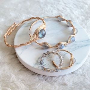 Peut inclure: Quatre bijoux : deux bracelets et deux bagues. Les bracelets et les bagues sont faits de fil doré et présentent des pierres précieuses irisées bleu-gris. Les bijoux sont présentés sur un plateau rond en marbre blanc.