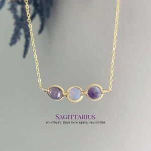 May include: Gold chain necklace with three round gemstone beads. The beads are amethyst, blue lace agate, and lepidolite. The text "SAGITTARIUS amethyst, blue lace agate, lepidolite" is below the necklace.