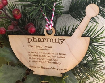 May include: A wooden ornament in the shape of a mortar and pestle. The ornament has the word "pharmily" and a definition printed on it. It is hanging from a red and white string, surrounded by greenery and red berries.