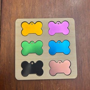 Könnte beinhalten: Sechs leere, hundeknochenförmige Metallanhänger auf einem Holzbrett. Die Anhänger sind in verschiedenen Farben: Gelb, Rosa, Grün, Blau, Schwarz und Roségold.