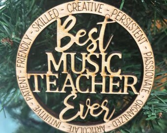 Puede incluir: Un adorno de madera con el texto "Best Music Teacher Ever" rodeado de un círculo de adjetivos positivos. El adorno está colgado de un árbol de Navidad.