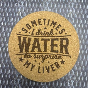 Puede incluir: Posavasos redondo de corcho con el texto "Sometimes I drink WATER to surprise MY LIVER" en letras negras. El posavasos es de color marrón claro y tiene una textura natural de corcho. El texto está dispuesto en forma circular.