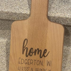 May include: A wooden chopping board with a handle, engraved with the words "home", "EDGERTON, WI", and the coordinates "42.8353° N. 89.0676° W".