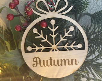 Op de afbeelding: Een houten kerstversiering in de vorm van een cirkel met een sneeuwvlokdesign en het woord "Herfst" erop. De versiering hangt aan een rood-wit koord.