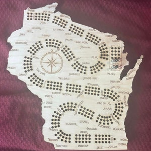 May include: A wooden game board shaped like the state of Wisconsin with the names of cities and towns throughout the state. The board has a compass in the center and a series of holes for playing pieces.