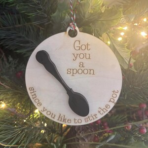 May include: A round wooden Christmas ornament with the text "Got you a spoon since you like to stir the pot" and a black spoon design. The ornament hangs from a red and white string.