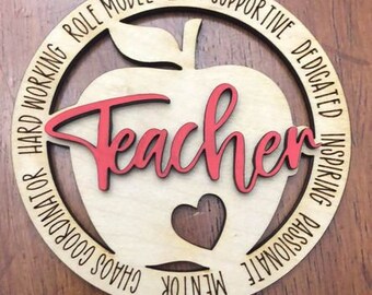 Può includere: Un ornamento di legno a forma di mela con la parola "Teacher" in lettere rosse all'interno. L'ornamento è circondato da un cerchio con le parole "Hard Working Role Model 2022 Supportive Dedicated Inspiring Passionate Mentor Chaos Coordinator" scritte intorno.