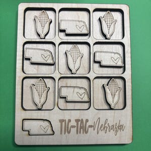 Puede incluir: Juego de Tic-Tac-Toe de madera con nueve casillas. Las piezas son mazorcas de maíz y el contorno de Nebraska con un corazón. Las palabras "TIC-TAC-Nebraska" están grabadas en el tablero.