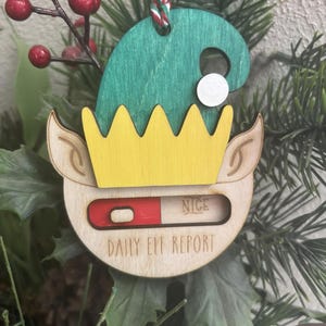 May include: Wooden elf ornament with a green hat, yellow trim, and a red and white slider that reads "NICE" and "DAILY ELF REPORT".
