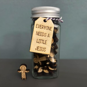 May include: A clear glass jar with a silver lid and a wooden tag that reads "EVERYONE NEEDS A LITTLE JESUS." The jar contains small, dark objects. A small wooden figure of Jesus stands in front of the jar.