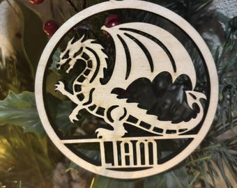 May include: A wooden Christmas ornament with a black silhouette of a dragon and the name "Liam" written below. The ornament is hanging on a Christmas tree.