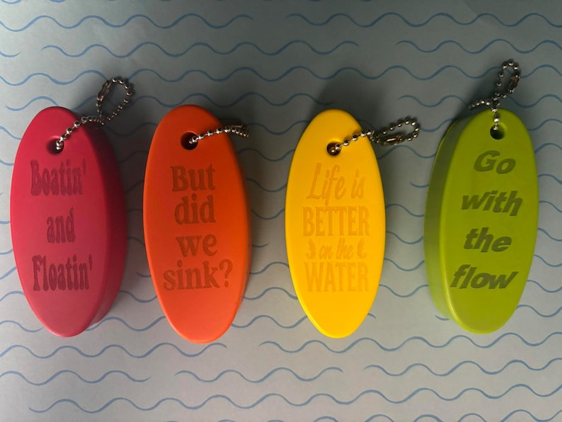 May include: Four oval-shaped keychains with different sayings. The keychains are red, orange, yellow, and green. The sayings are "Boatin' and Floatin'," "But did we sink?", "Life is better on the water", and "Go with the flow."