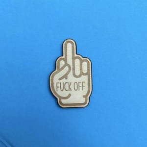 May include: A wooden cutout of a hand giving the middle finger, with the words "FUCK OFF" engraved on it. The cutout is light brown with a darker brown outline, set against a blue background.