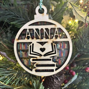 Puede incluir: Adorno navideño de madera con el nombre "ANNA" recortado. El adorno presenta un diseño de estantería visible a través de los recortes, con una pila de libros y un corazón. Cuelga de una cuerda verde y blanca.