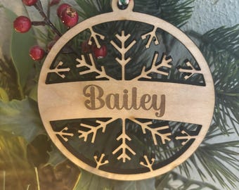 Op de afbeelding: Een houten kerstversiering met een sneeuwvlokdesign en de naam "Bailey" erop. De versiering hangt aan een groene kerstboomtak.