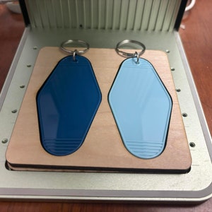 May include: Two keychains with blue and light blue motel key-shaped tags on a wooden base. The keychains are attached to silver key rings.