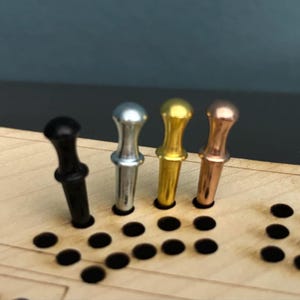 Könnte beinhalten: Nahaufnahme eines hölzernen Cribbage-Bretts mit vier Metallstiften. Die Stifte sind schwarz, silber, gold und kupferfarben. Das Brett hat Reihen von Löchern zum Zählen.