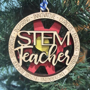 Puede incluir: Adorno de madera con las palabras "STEM Teacher" en un círculo. El círculo está rodeado de palabras que describen a un profesor de STEM, como "inspirador", "innovador", "curioso", "atractivo", "inventivo", "alentador", "constructor del futuro" y "de apoyo".