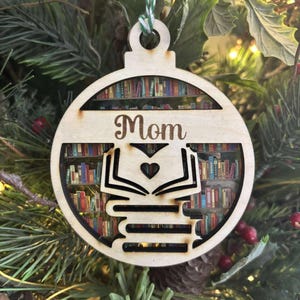 Puede incluir: Adorno navideño de madera en forma de bola, con la palabra "Mom" y una pila de libros con un libro abierto y un corazón recortado. El adorno tiene una escena de biblioteca en el interior y está colgado de un árbol de Navidad.