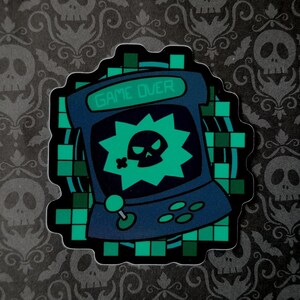 May include: A sticker with a retro arcade game design, featuring a skull and crossbones graphic. The screen displays "GAME OVER" in green. The sticker has a dark blue and green colour scheme, with a pixelated border.