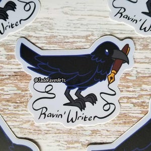 May include: A sticker featuring a black raven with blue accents, holding a brown pen. The sticker has the text "Ravin' Writer" in a stylized font, with the artist's name, "@JadeRavenArts".